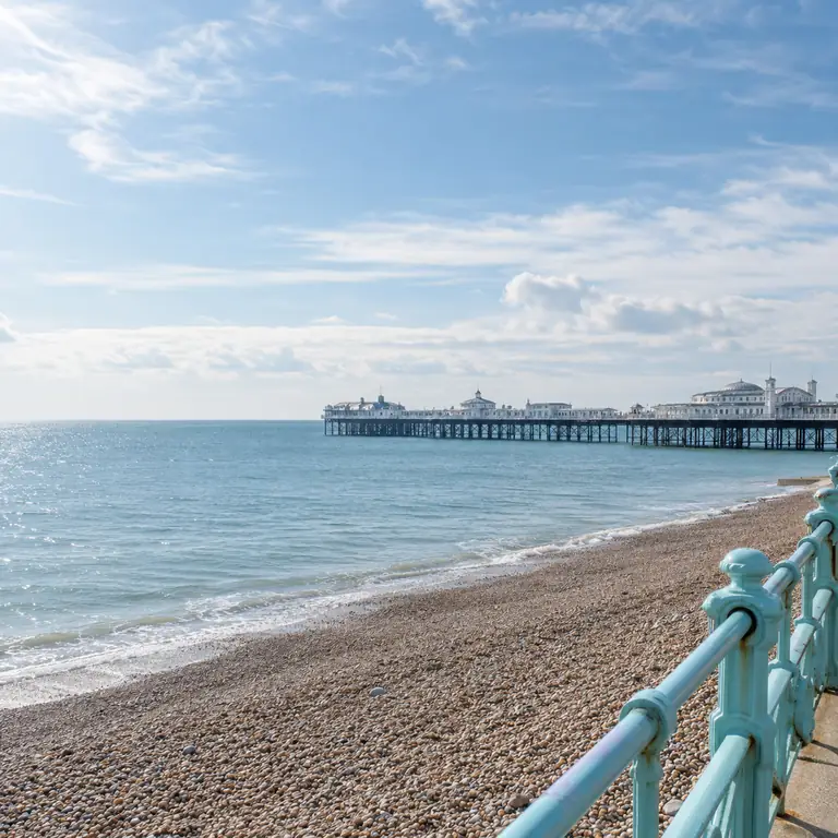 Açık mavi korkuluklar Brighton’ın çakıllı plajını çevrelerken iskele sakin denizin üzerinde uzanıyor.